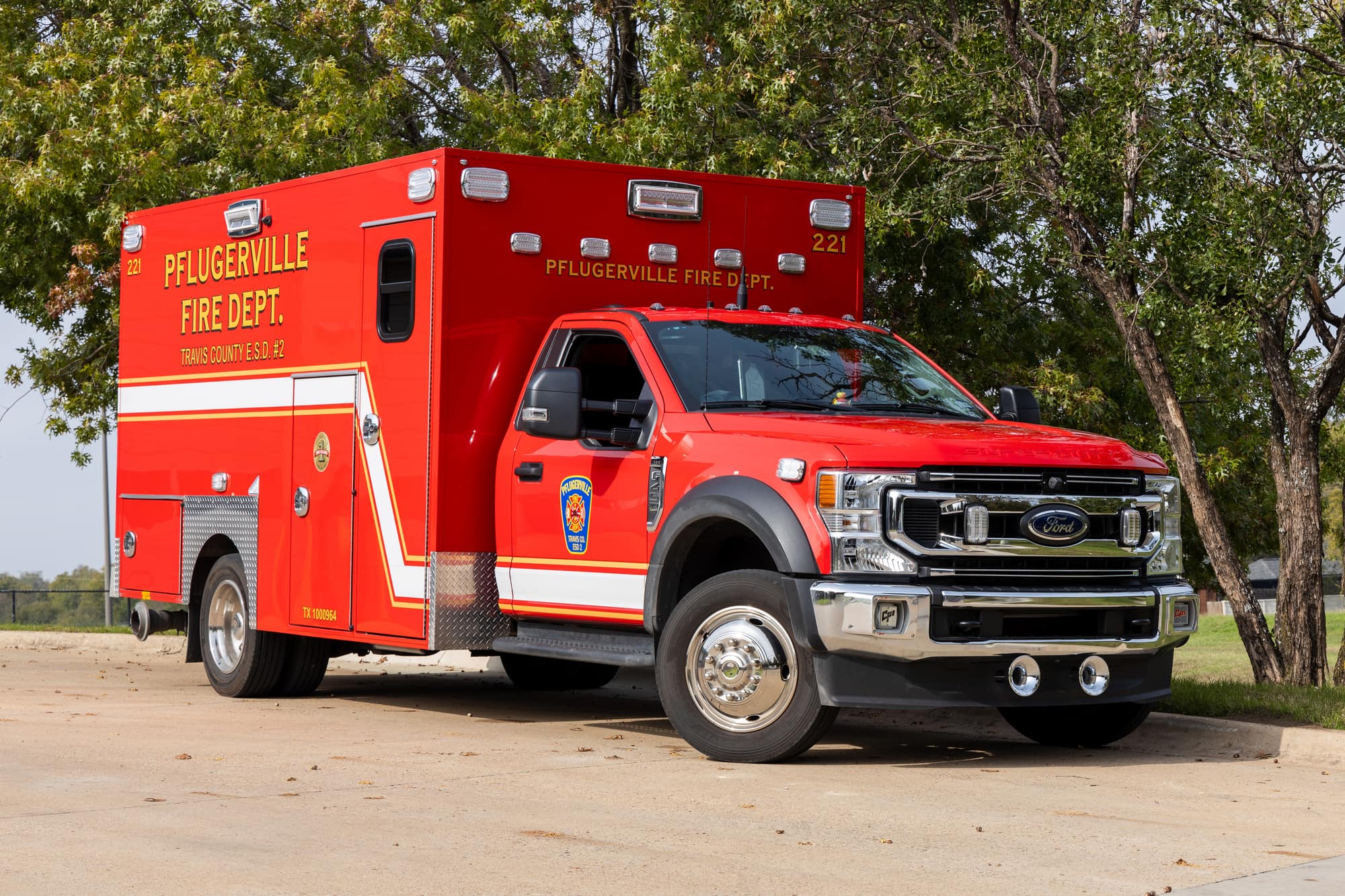 Red Pflugerville Fire Department Medic 221 ambulance parked along a curb near trees.