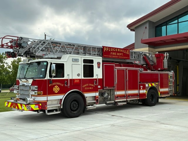 Red and white Pflugerville Fire Department Reserve Quint 201 aerial ladder truck parked outside.